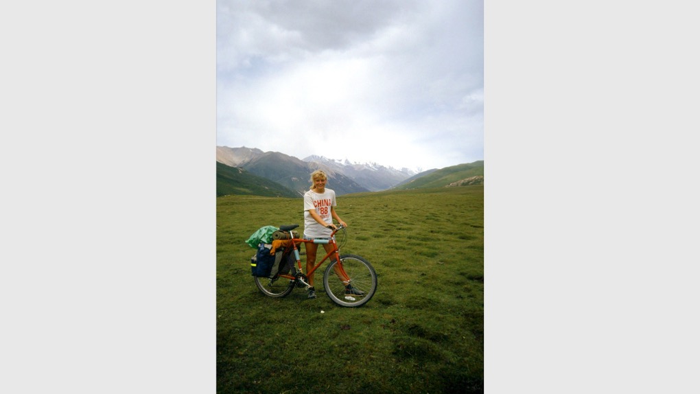 The image shows Karen stood on grass next to and holding a bicycle. There are mountains and clouds in the sky behind her and her belongings are on the back of the bike. She's holding the handlebars and smiling at the camera.