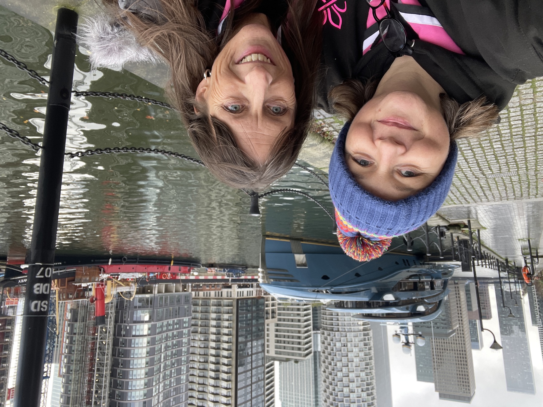 Two women are stood on pavement with a river next to them on the left. There are two boats in the background on the water and skyscrapers the other side of the river. The two women are stood close together, Emma on the right wearing a hot pink, black and white Scouts necker with a blue beanie hat on, and Teresa is on the left wearing a coat with a fluffy hood. They're both smiling at the camera