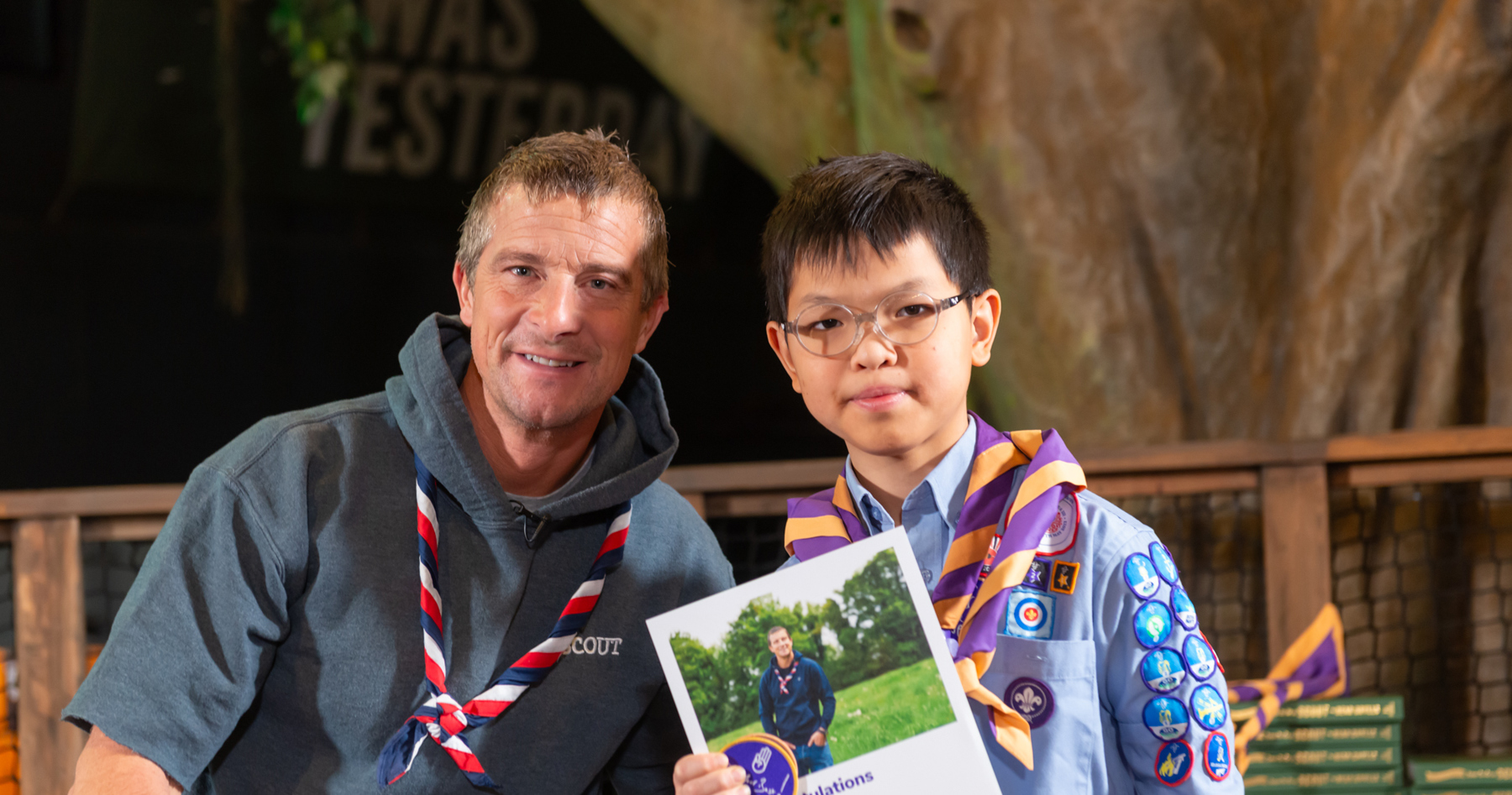 The image shows Chief Scout Bear Grylls stood with 2023 Unsung Hero award recipient, Ernest. Both are smiling at the camera, with Bear wearing a red, white and navy necker. Ernest is wearing glasses and his Scout shirt with badges on, as well as a purple and orange necker. Ernest is holding a certificate and a Never Give Up badge in his right hand.