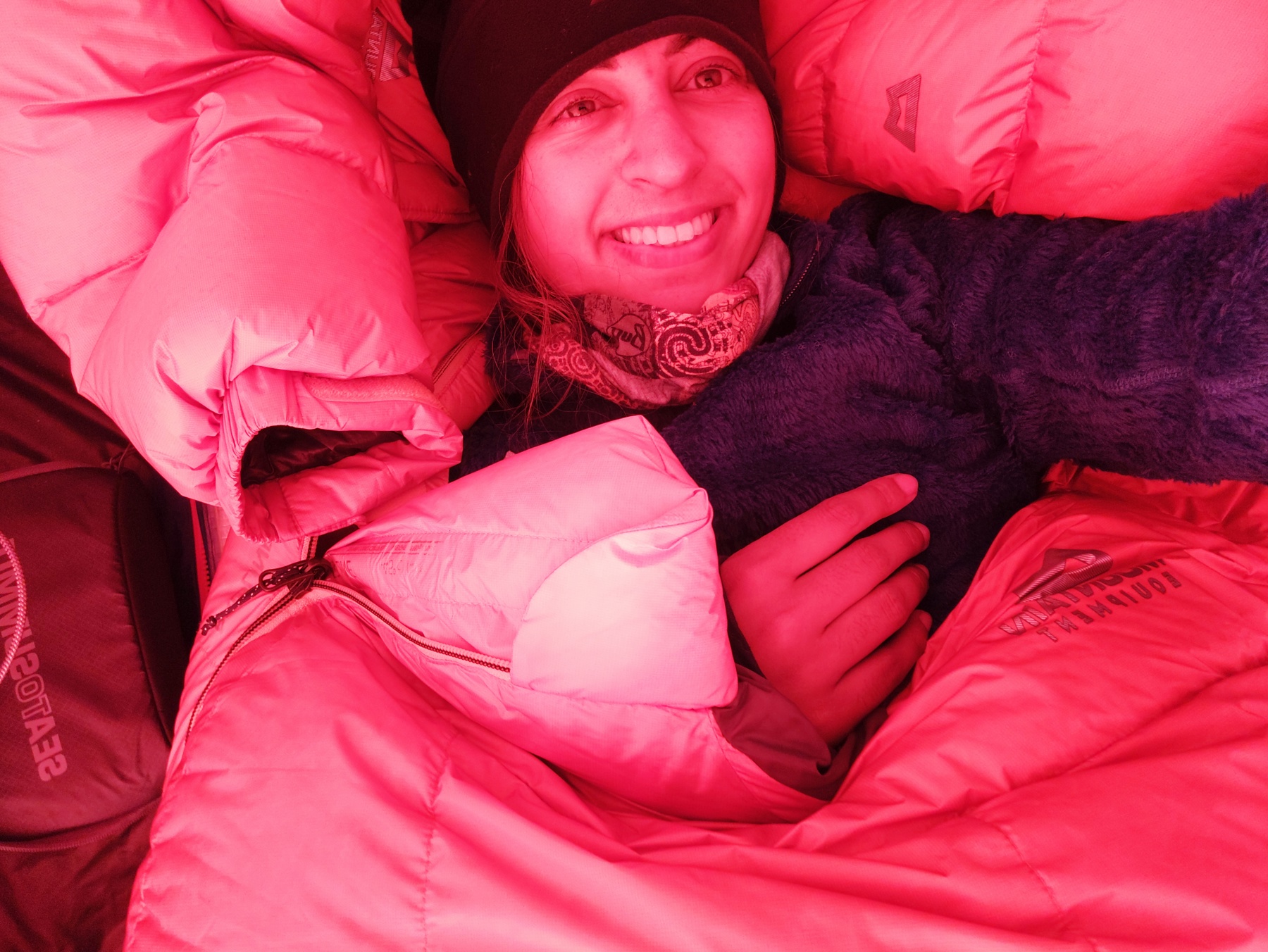 The image shows Preet Chandi MBE wearing a hat, neck warmer and fluffy jumper while she holds the camera above her head to take a selfie. She's smiling at the camera and she's wrapped up in a red sleeping bag.