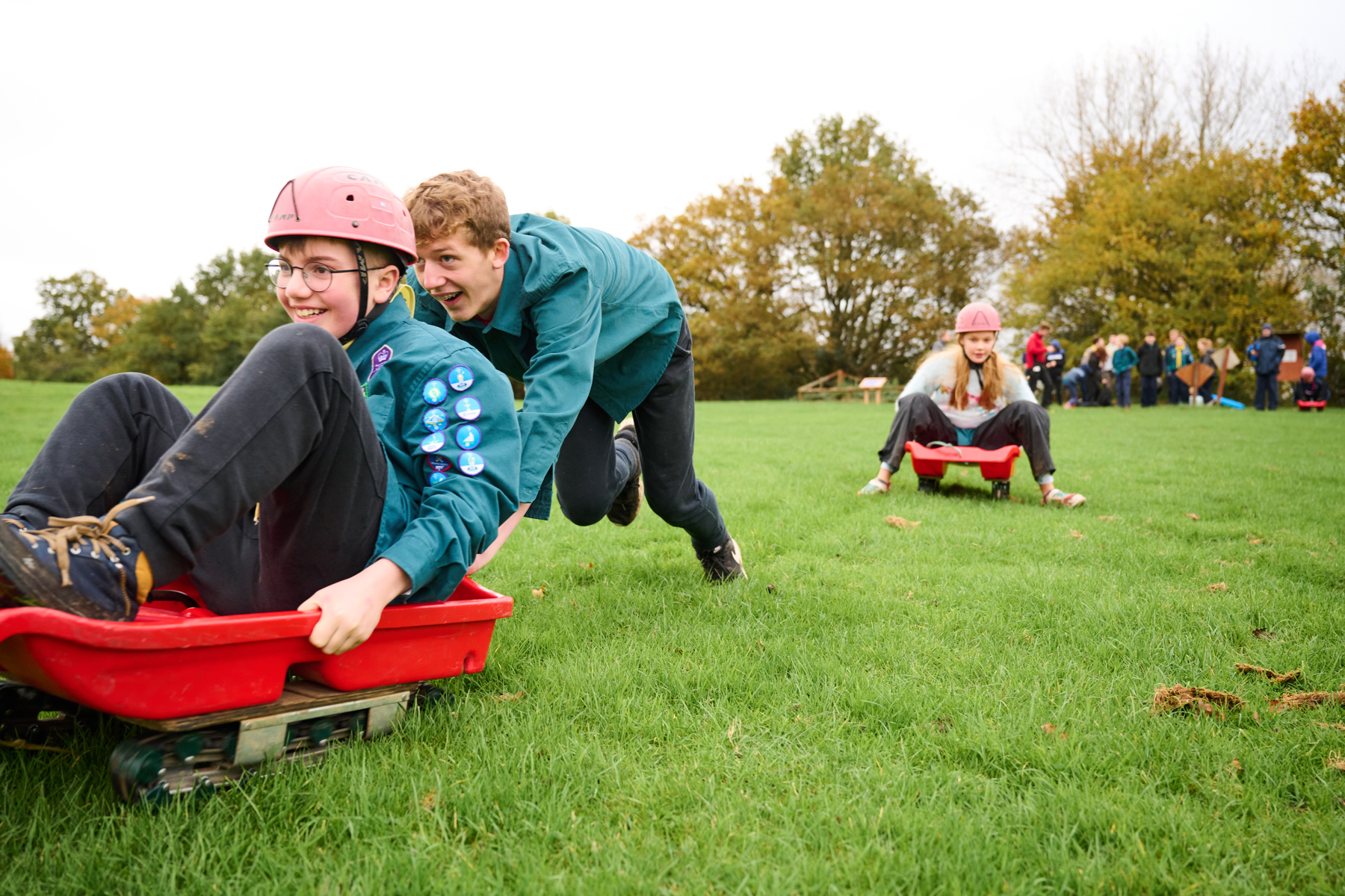 To the left, a Scout is sat on a red sledge wearing uniform and a pink helmet. Another Scout is pushing them down a grassy hill, leaning forwards. To the right, there's another Scout on a red sledge in the background, wearing a grey jumper and trousers, and a pink helmet. There are trees in the background and more young people at the top of the hill.